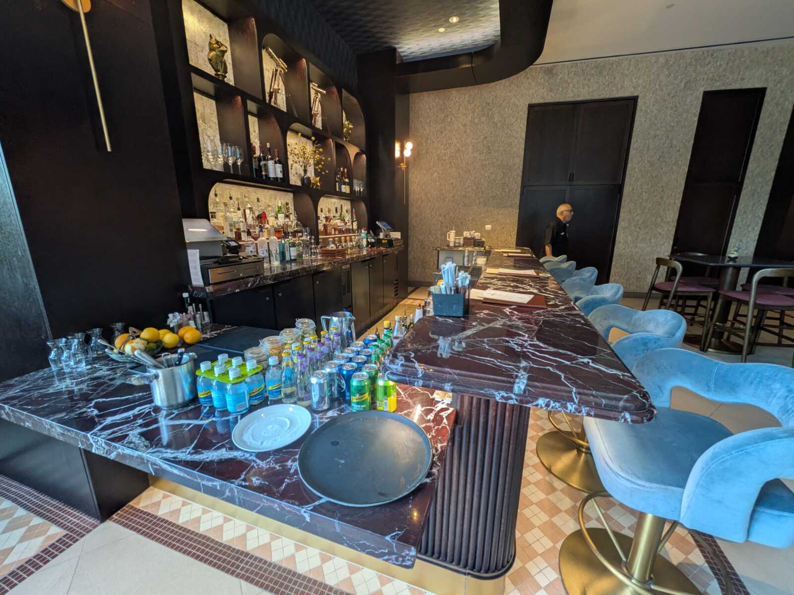 Bar with dark marble counter and turquoise velvet stools, liquor shelves along the back wall, and a bartender in the distance.