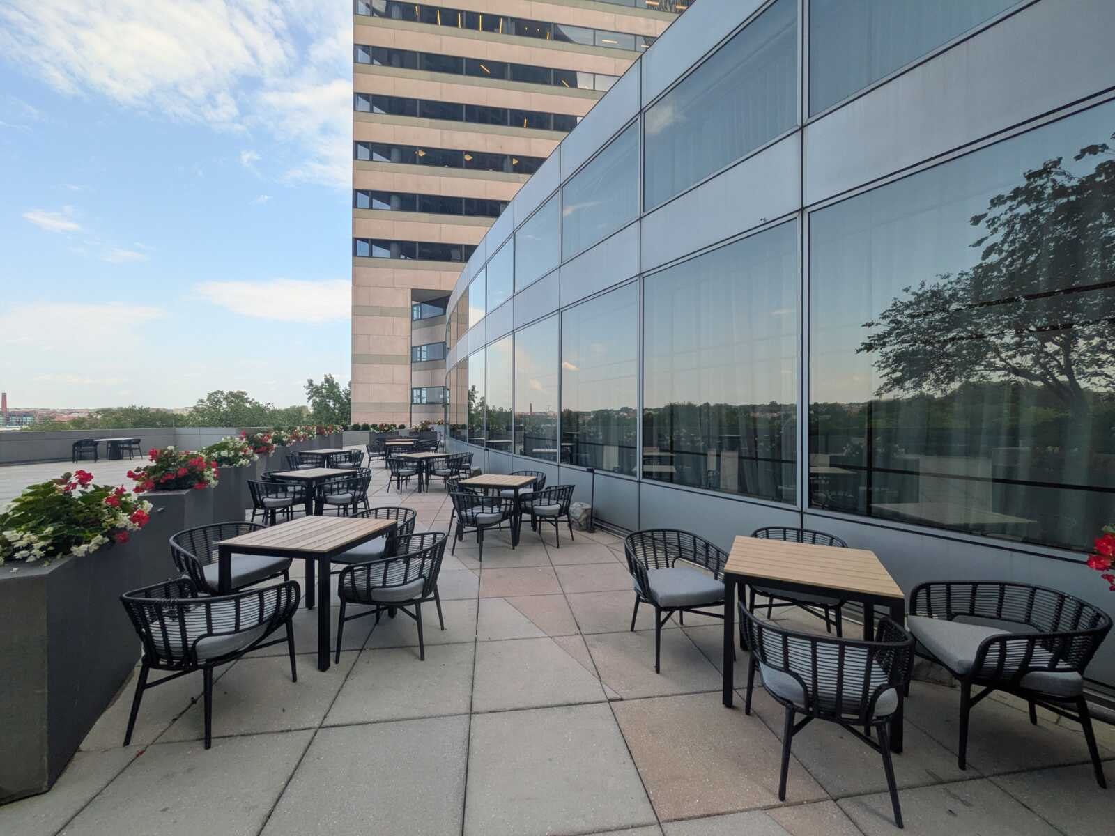 Rooftop patio with wooden tables and black chairs, flower-filled planters along the edge, beside a glass-walled building.