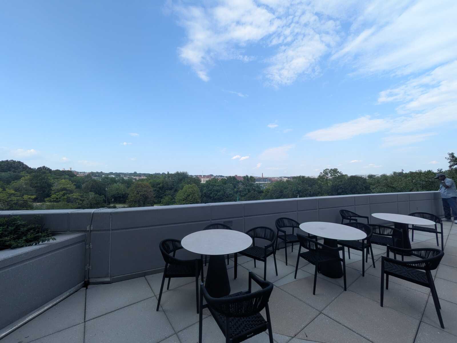 Rooftop terrace with round white tables and black chairs overlooking a tree-filled landscape under a blue, cloudy sky.