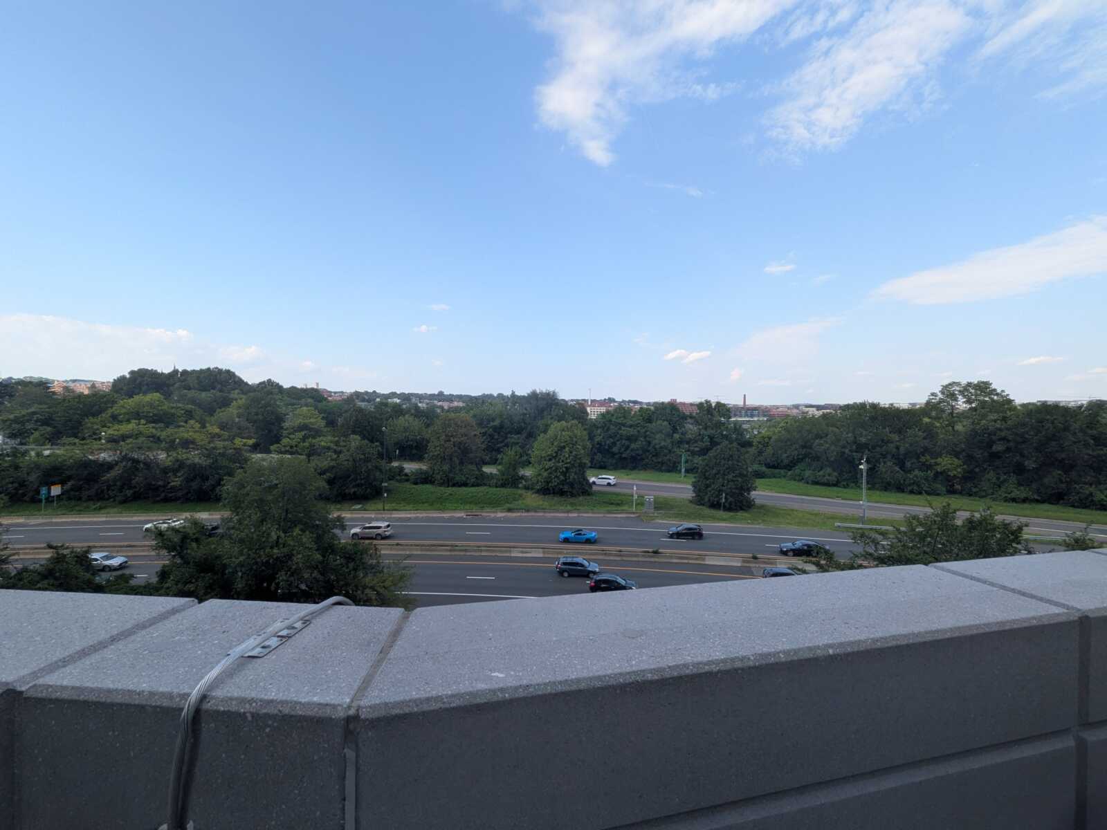 Rooftop edge in the foreground with a multi-lane highway below, lined by trees and a distant city skyline under a blue sky.