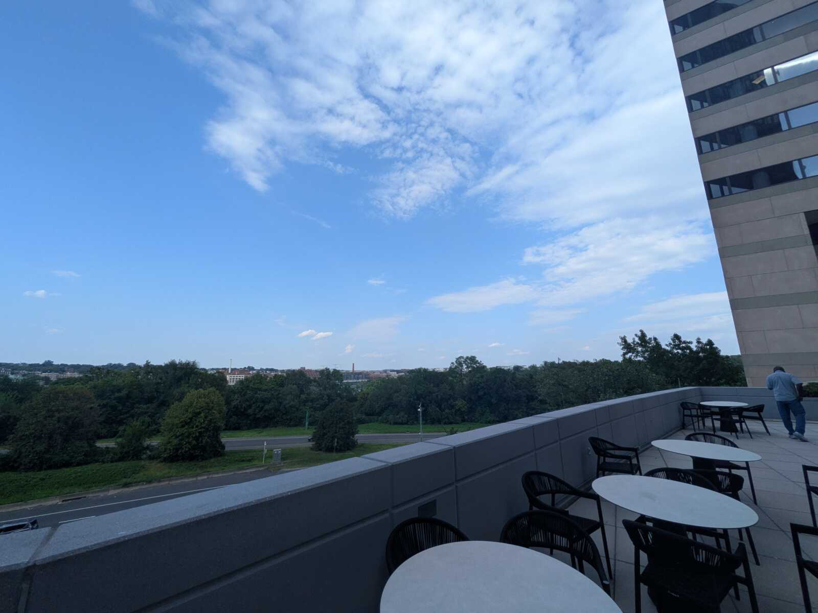 Rooftop terrace with round white tables and black chairs along a railing, a person standing near the edge, with a tall building on the right and a tree-filled landscape below under a blue, partly cloudy sky.