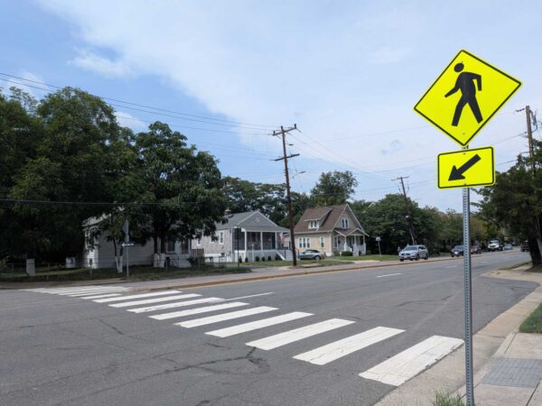 Urban intersection with a zebra crosswalk and bright yellow pedestrian sign; houses and trees in the background under a blue sky.