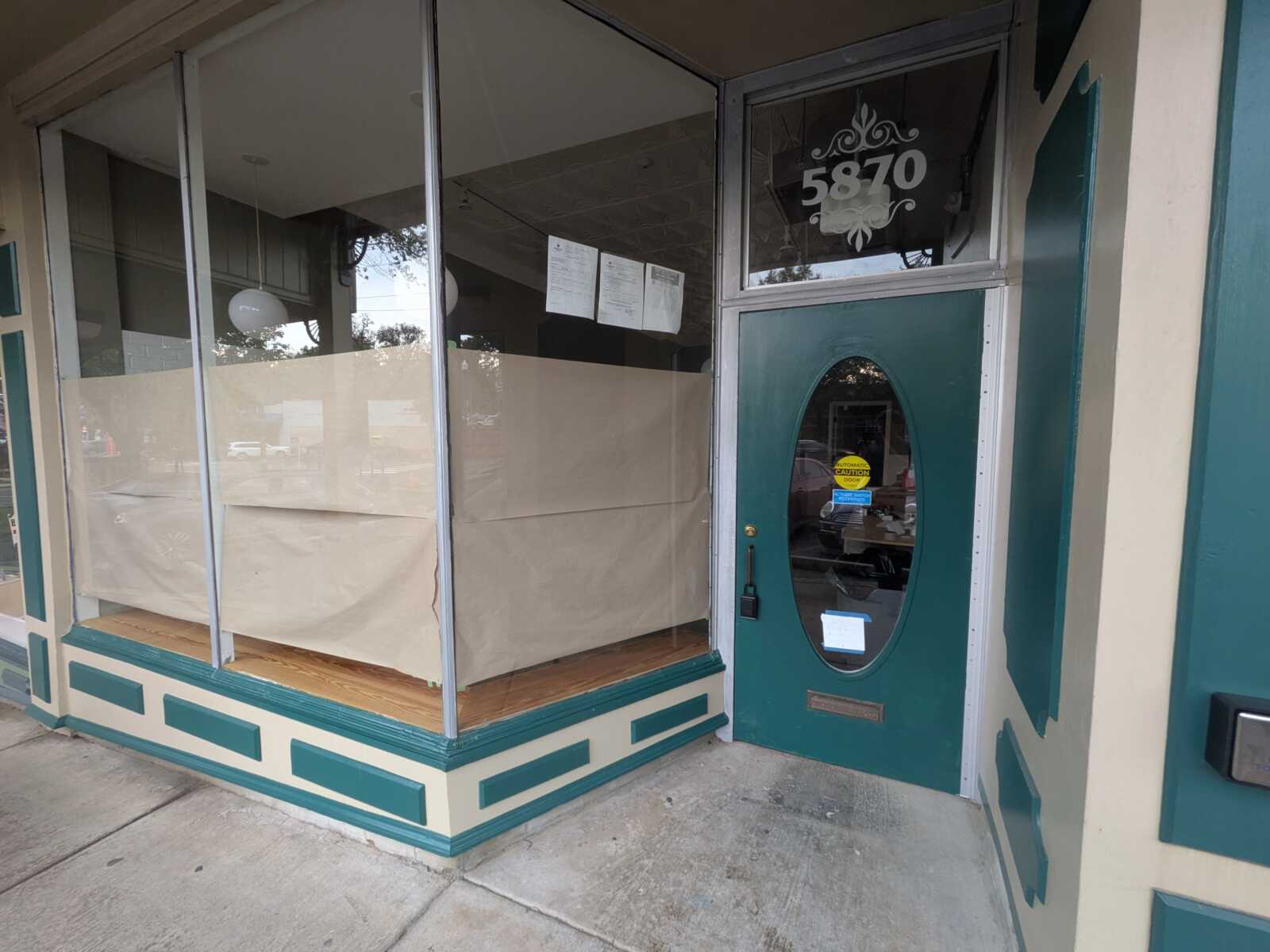 Vacant storefront with large glass windows covered by beige paper, teal trim, and a teal door marked 5870.