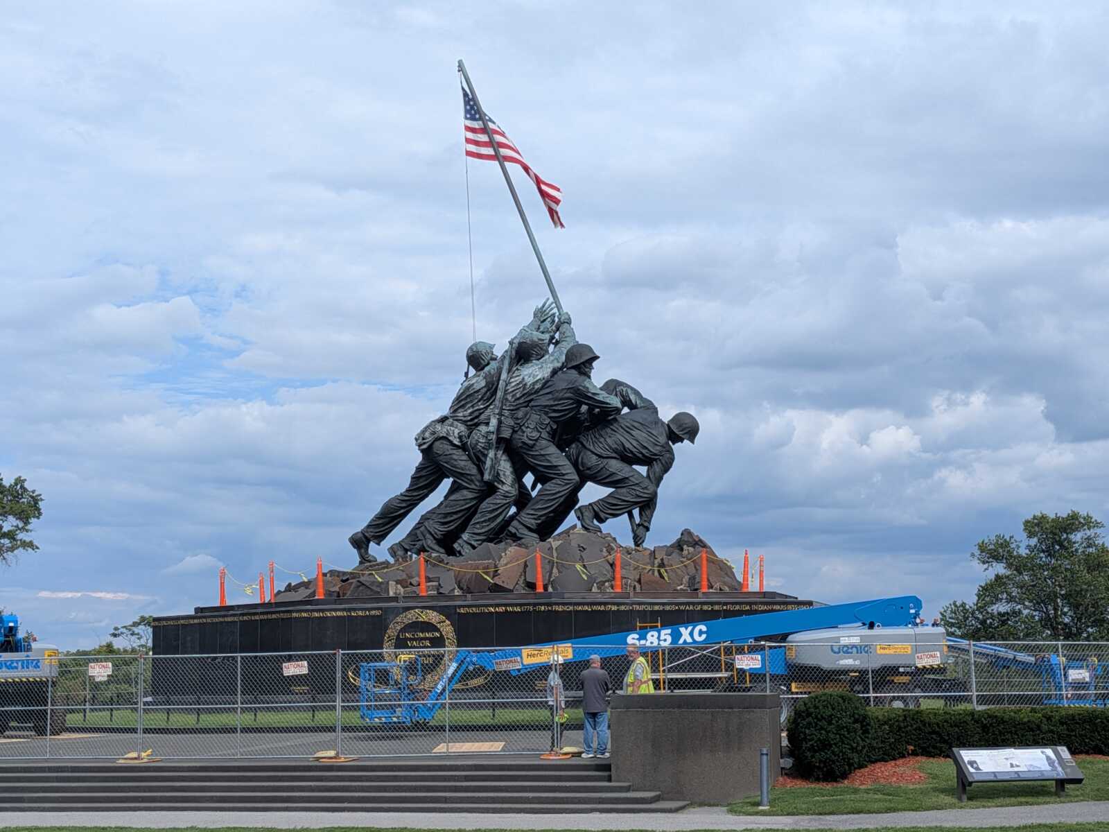 Bronze sculpture of soldiers raising an American flag atop a rocky pedestal at a war memorial, with fencing and construction equipment in the foreground.