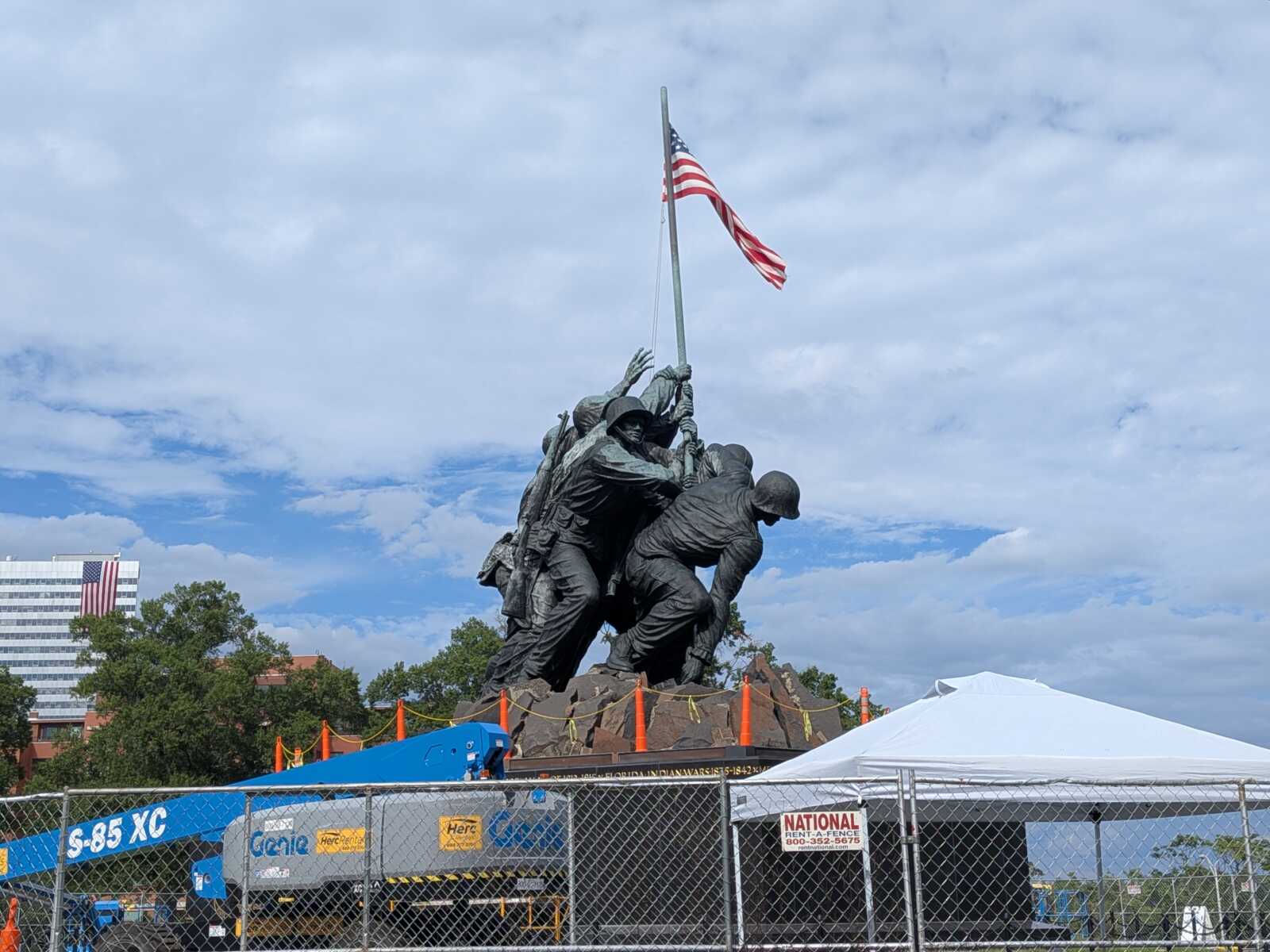 Bronze sculpture of soldiers raising an American flag on a tall pole, with a blue sky and construction fencing nearby.