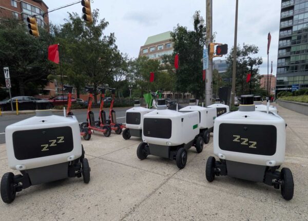 White autonomous delivery robots with black front screens and four wheels parked on a city sidewalk. In the background, orange scooters and buildings line the street.