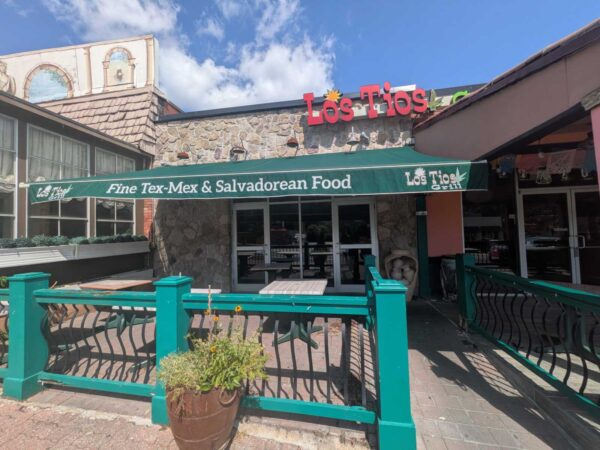Exterior of Los Tios restaurant with a green awning reading 'Fine Tex-Mex & Salvadoran Food' and teal fencing against a stone façade under a blue sky.