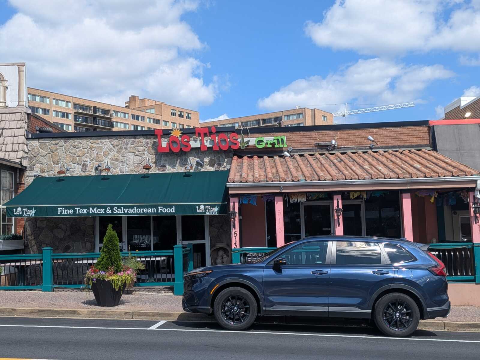 Exterior of Los Tios Grill, a Tex‑Mex and Salvadorian restaurant with a stone façade and green awning, and a blue SUV parked in front.