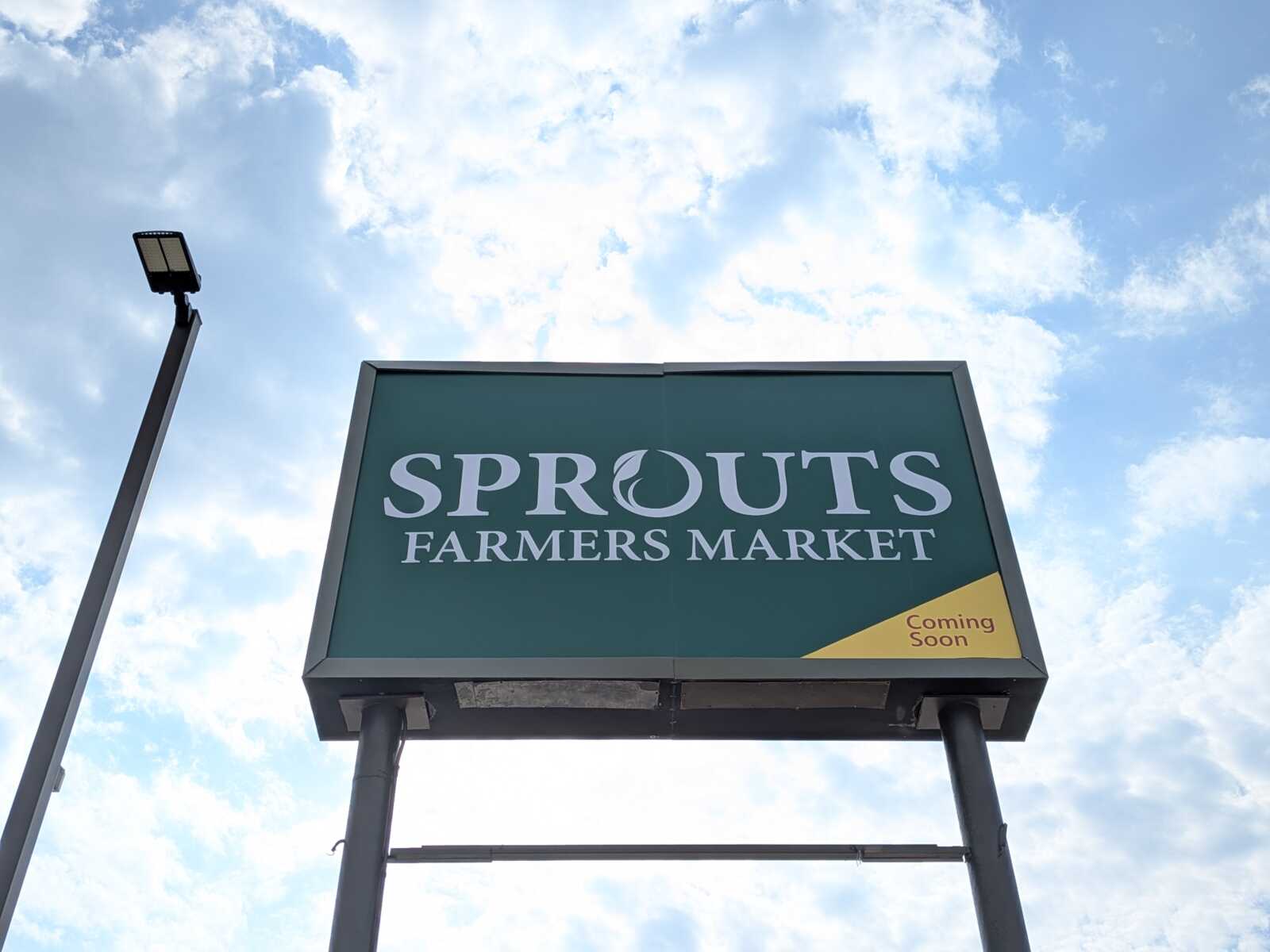 Green Sprouts Farmers Market sign against a blue sky with a yellow 'Coming Soon' tag in the bottom-right corner.