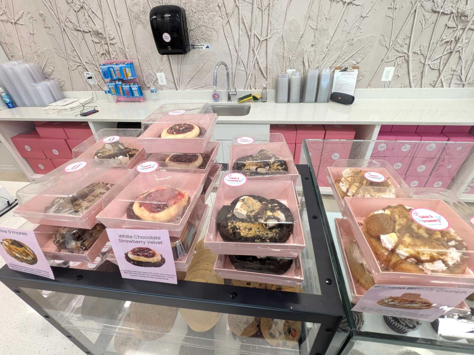 Assorted donuts in pink display boxes on a bakery counter, labels show flavors like White Chocolate Strawberry Velvet with a glass display case around them.