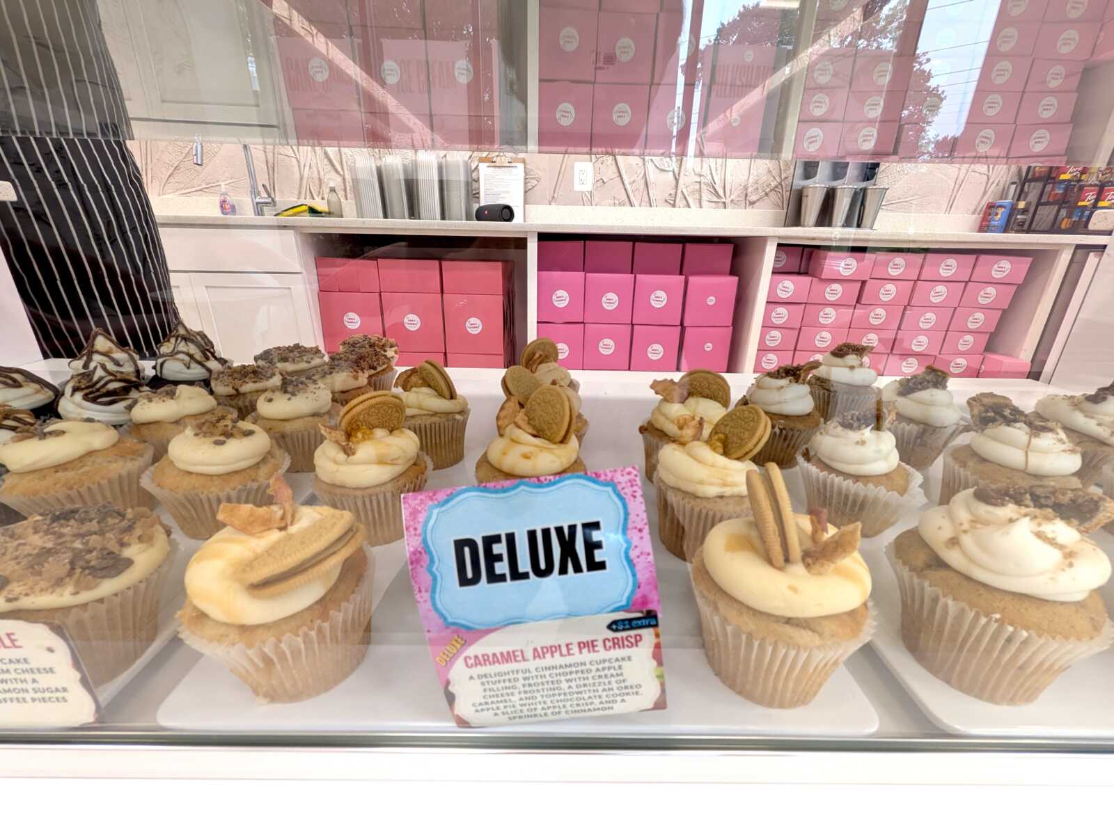 Assorted cupcakes with swirled frosting and toppings in a glass bakery display; a pink 'DELuxe' sign and product card are in front.