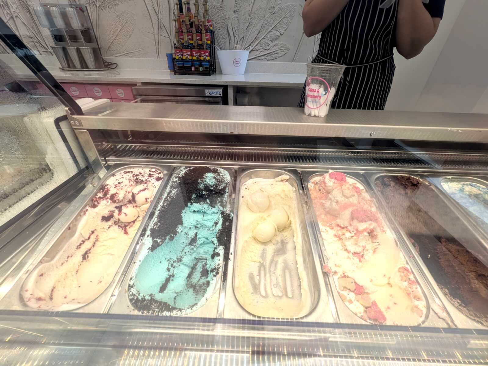 Ice cream shop display with multiple flavors in metal tubs; a staff member in a striped apron stands behind the counter.