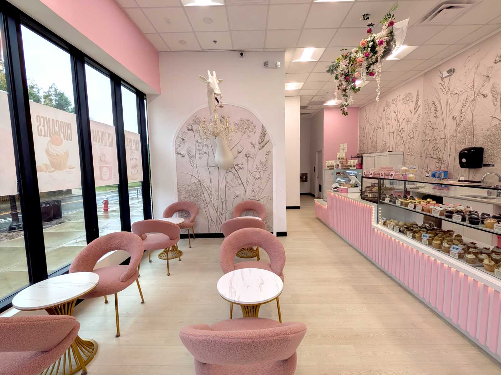 Pastel pink bakery seating area with round marble tables and pink upholstered chairs beside a glass pastry display case.