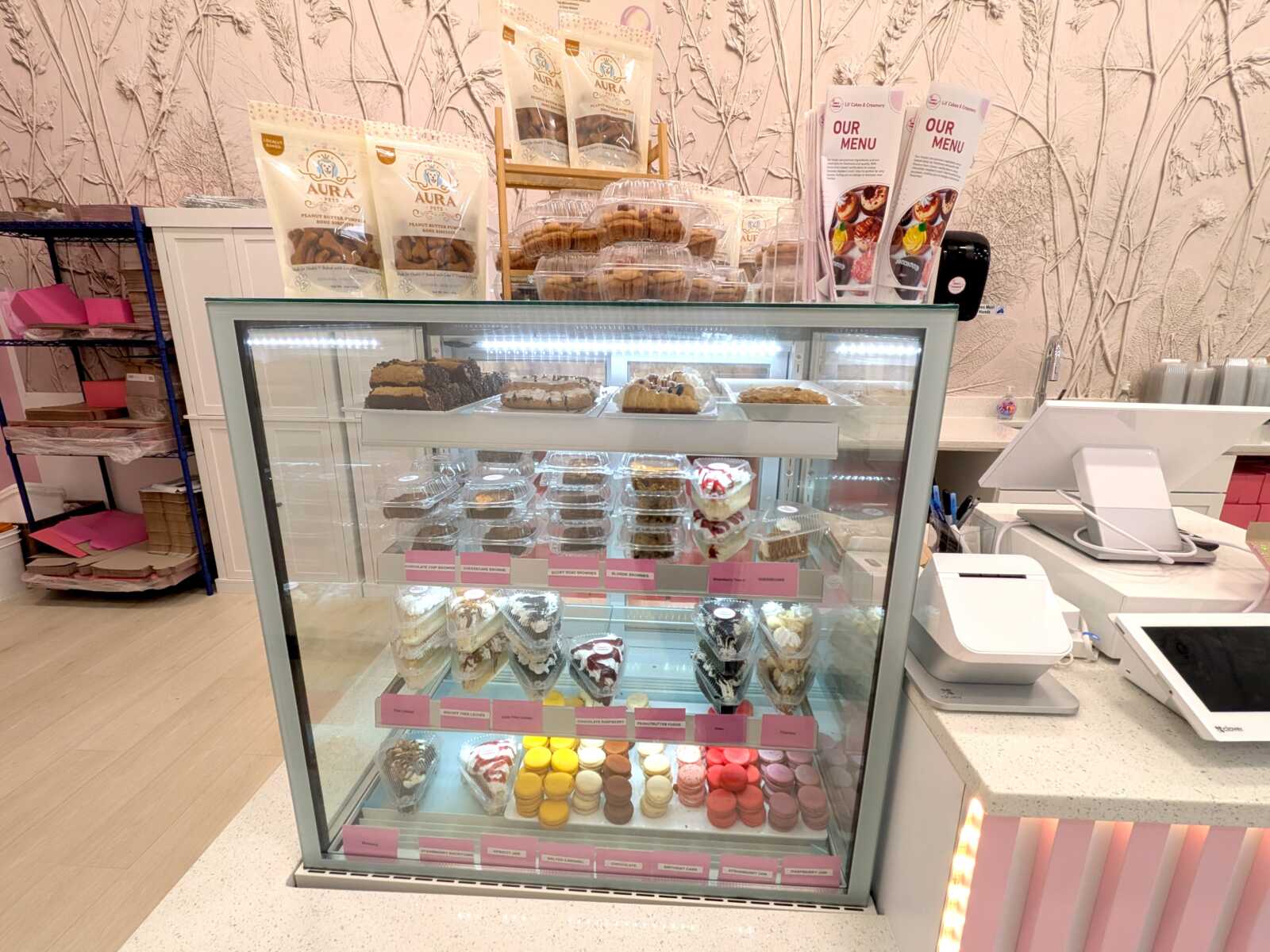 Glass-front bakery display case with assorted cakes, pastries, and macarons on multiple shelves in a pink-themed shop.