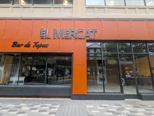 Storefront of El Mercat tapas bar with orange panels and bold metal sign reading 'EL MERCAT' and 'Bar de Tapas' in cursive, glass doors and windows behind