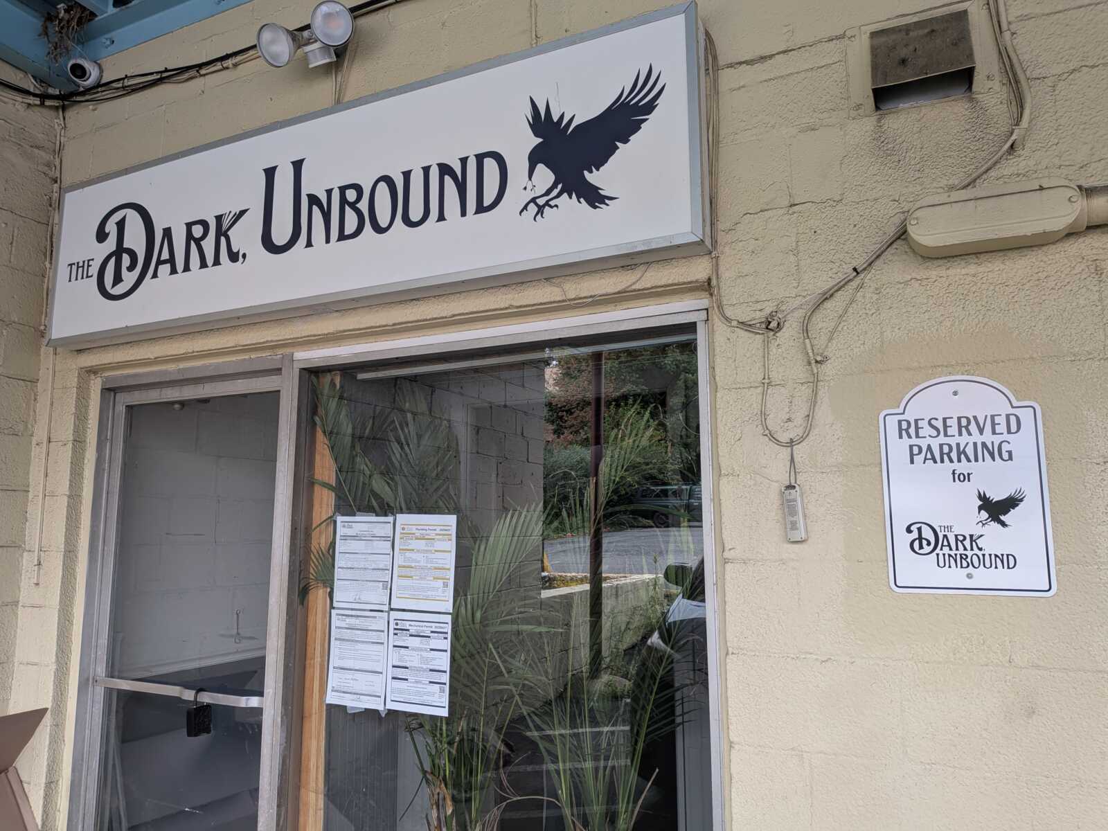 Storefront sign reading 'The Dark Unbound' with a flying crow silhouette above a glass door and a reserved parking sign on the wall