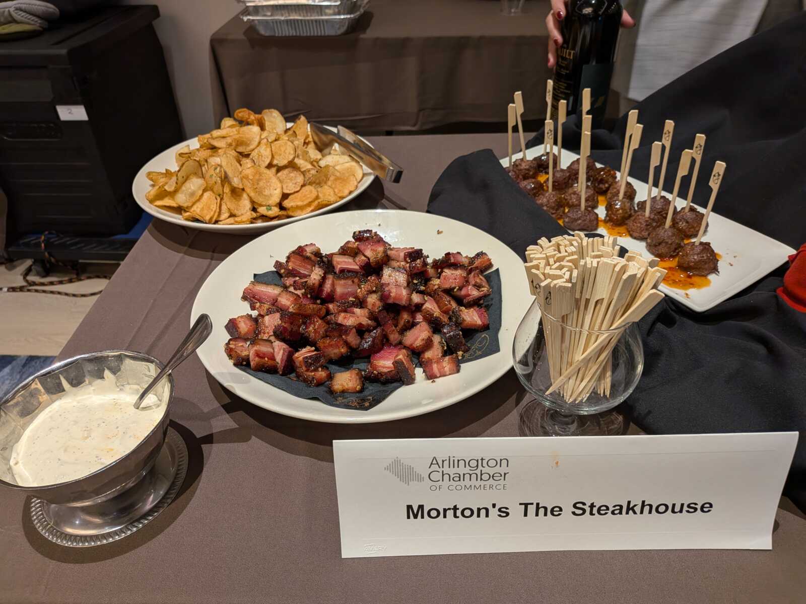 Plate of cubed, pepper-crusted steak bites with a side of potato chips and meatball skewers; Arlington Chamber of Commerce sign in foreground.