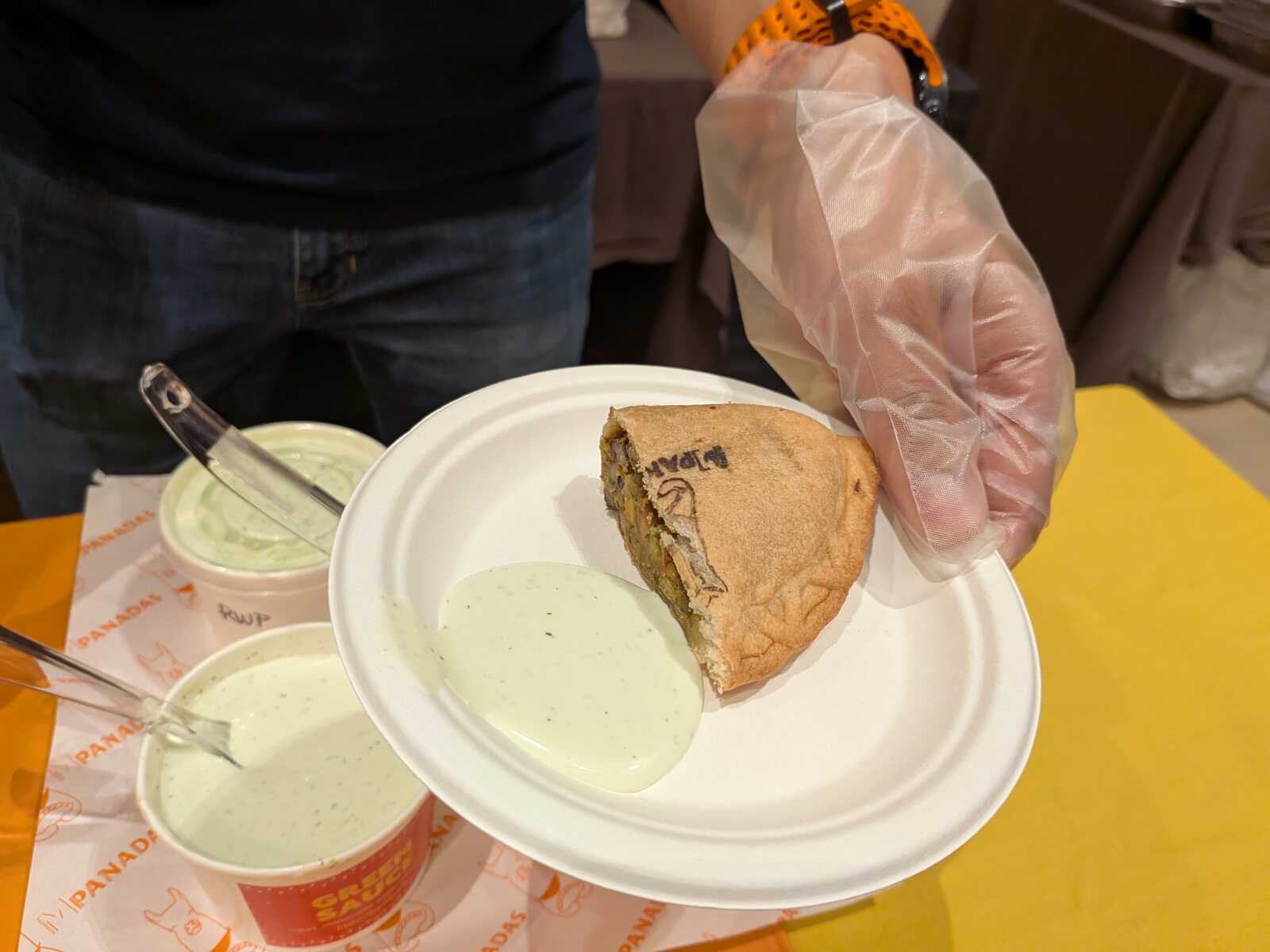 Gloved hand holding a white plate with a slice of pie and green dipping sauce; two cups of green sauce on an orange-drapped counter nearby.