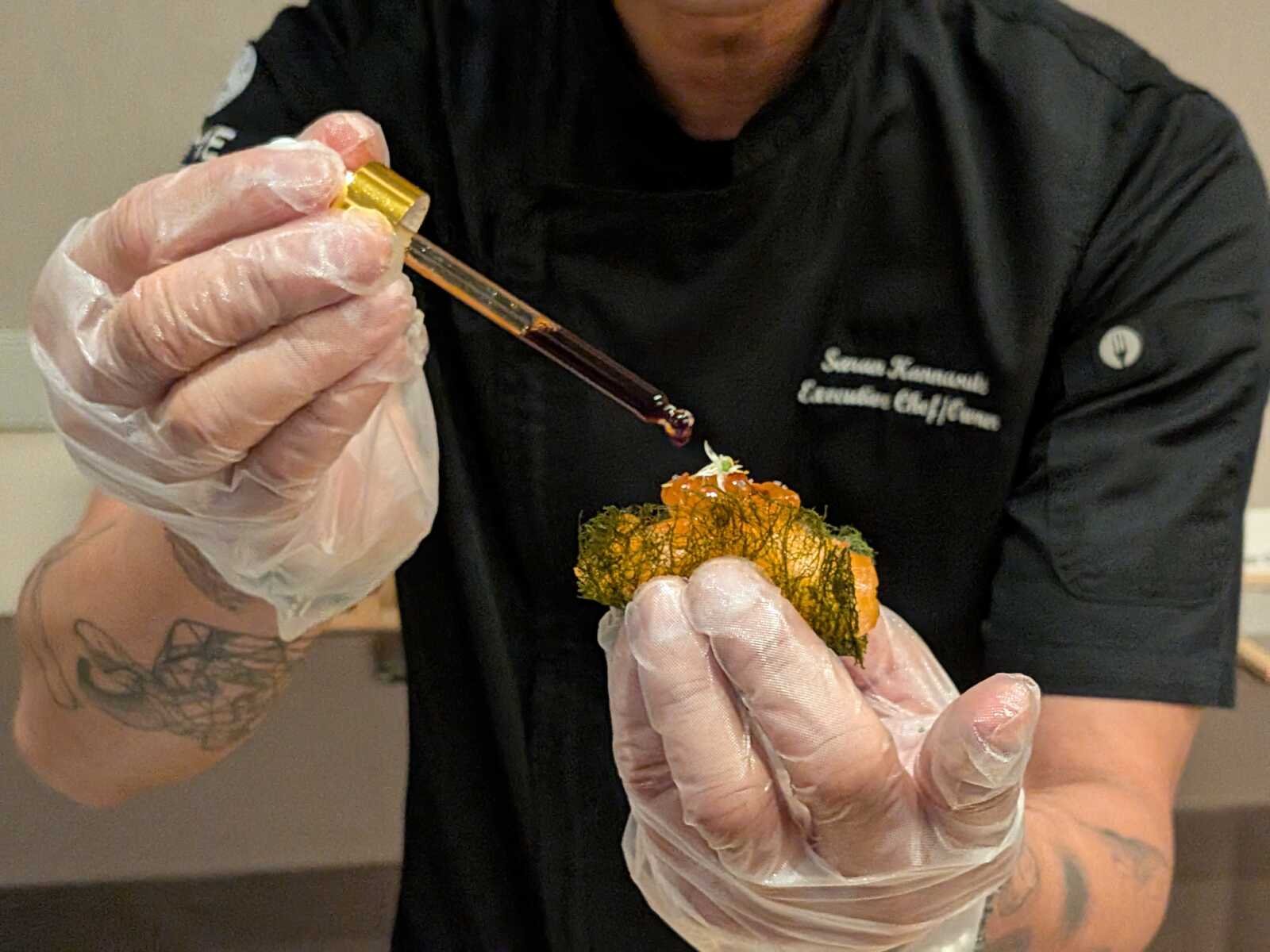 Chef in black uniform wearing gloves drips sauce from a dropper onto a plated dish.