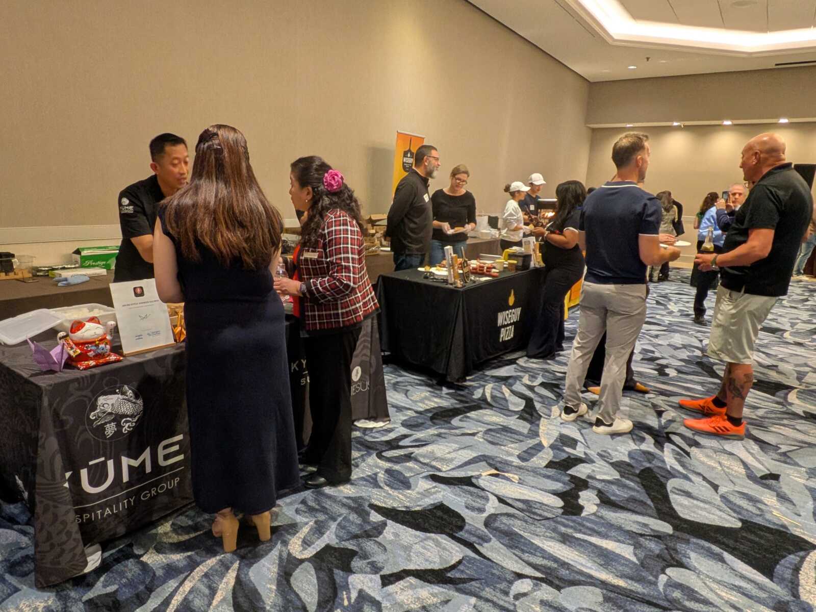 Attendees converse at vendor booths set up along a conference-room hall during a trade show or expo session, with products on display and staff assisting visitors or answering questions.