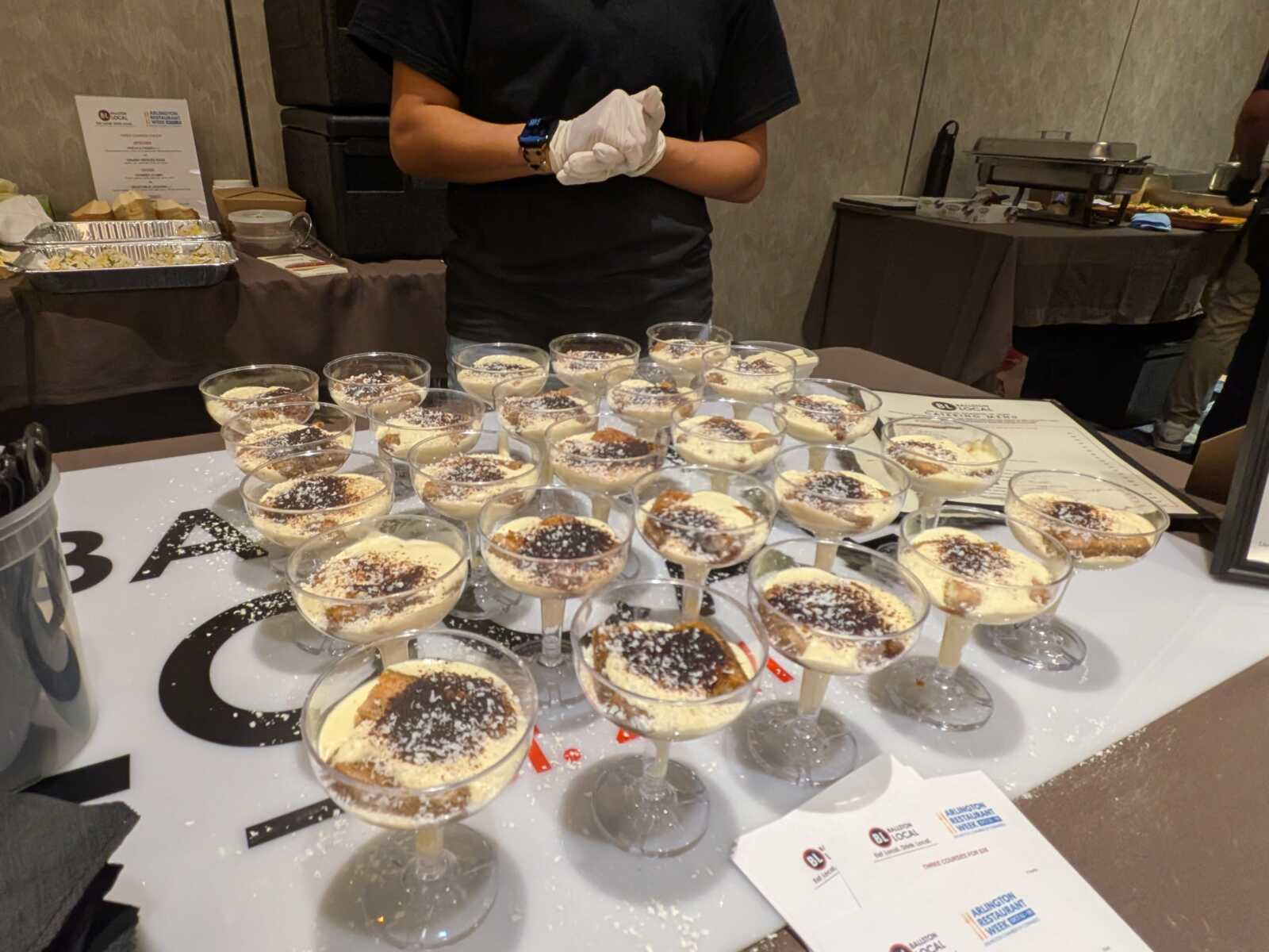 Assorted dessert cups arranged on a white table at a catered event, with a staff member in black gloves behind.