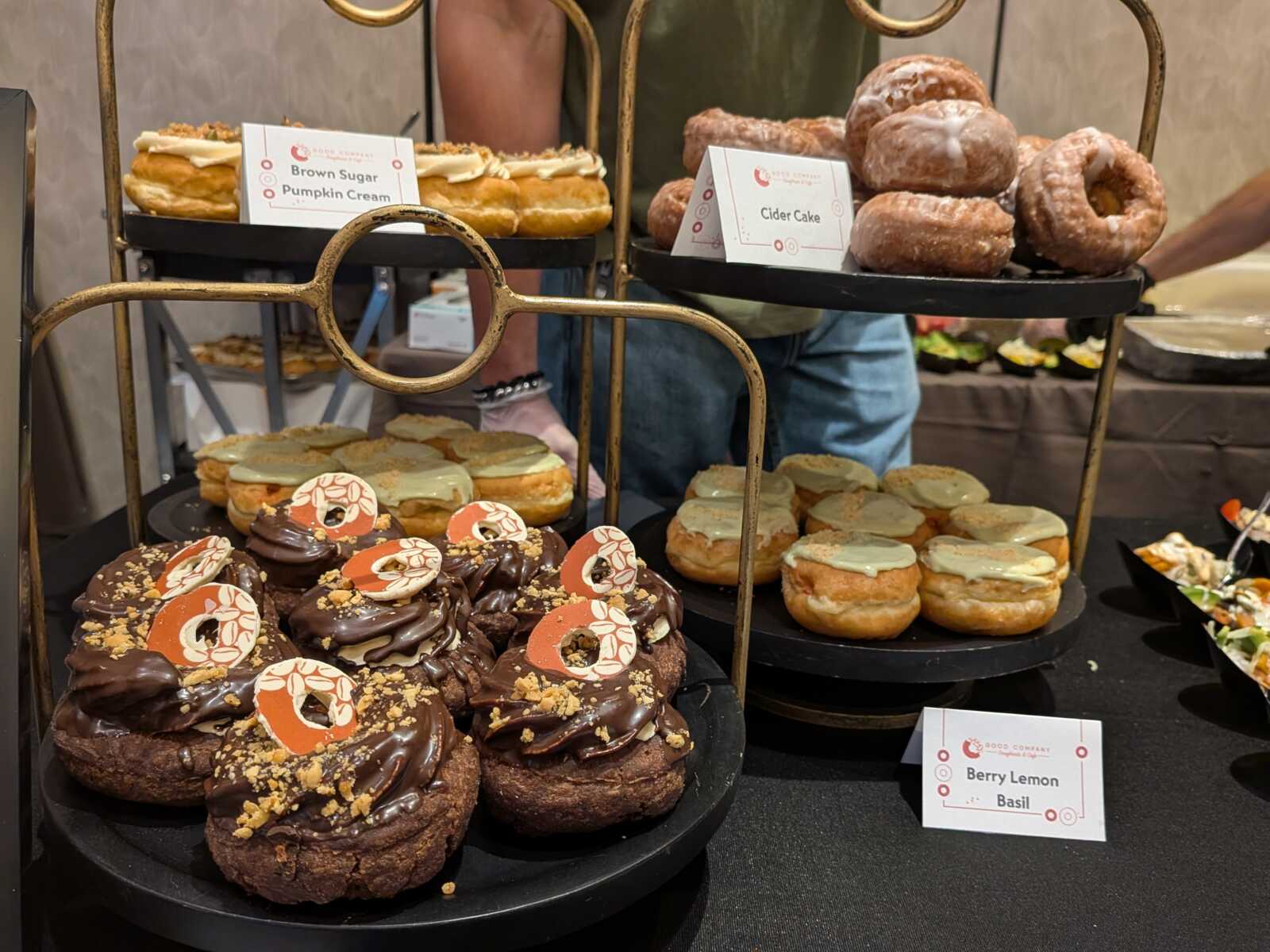 Assorted donuts on a curved, gold-toned display stand in a bakery, with chocolate-glazed donuts in the foreground and other flavors on the upper tier.
