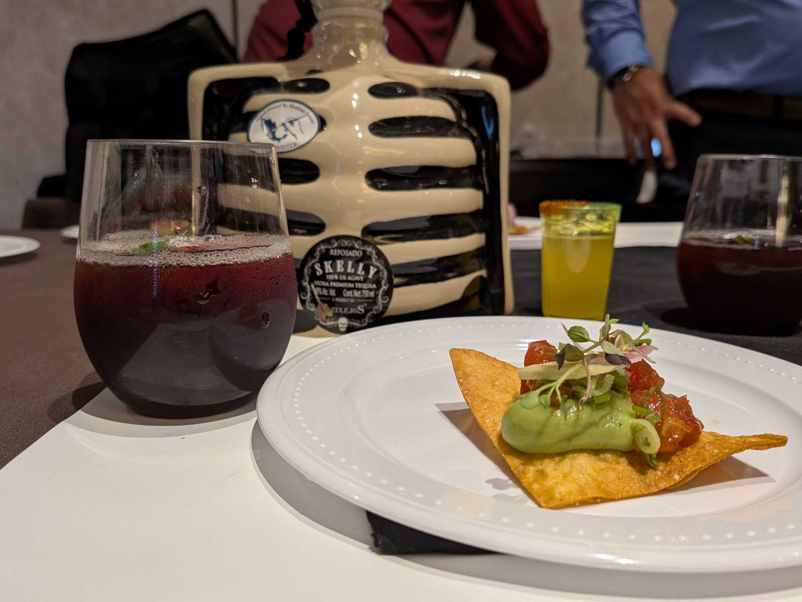 Glass of dark red beverage on a white table, foregrounded by a plate with avocado-topped tortilla chip and salsa, decorative bottle in background.
