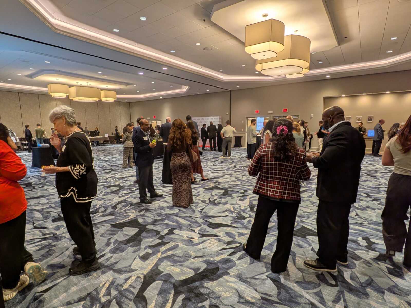 People mingle and chat with drinks at a large indoor conference reception in a ballroom with patterned carpet and hanging light fixtures.