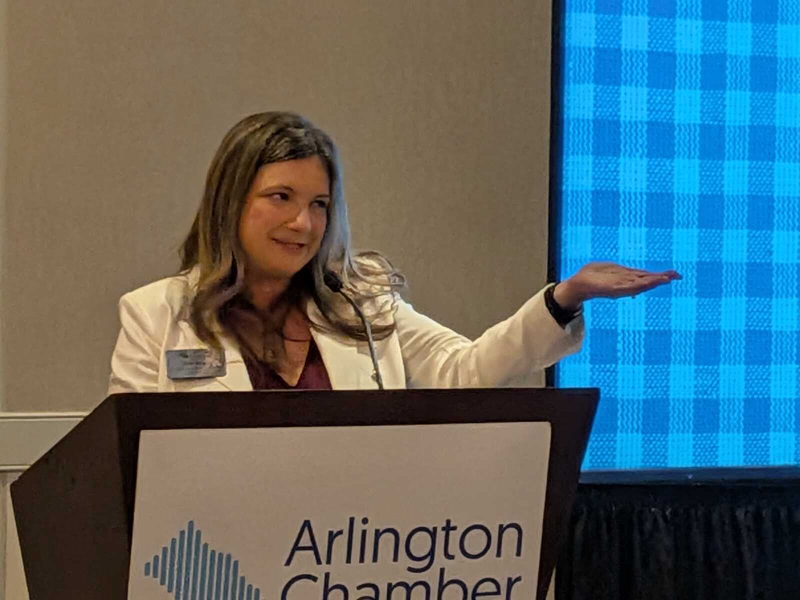Woman in a white blazer speaks at a podium for an Arlington Chamber event, gesturing with her right hand.