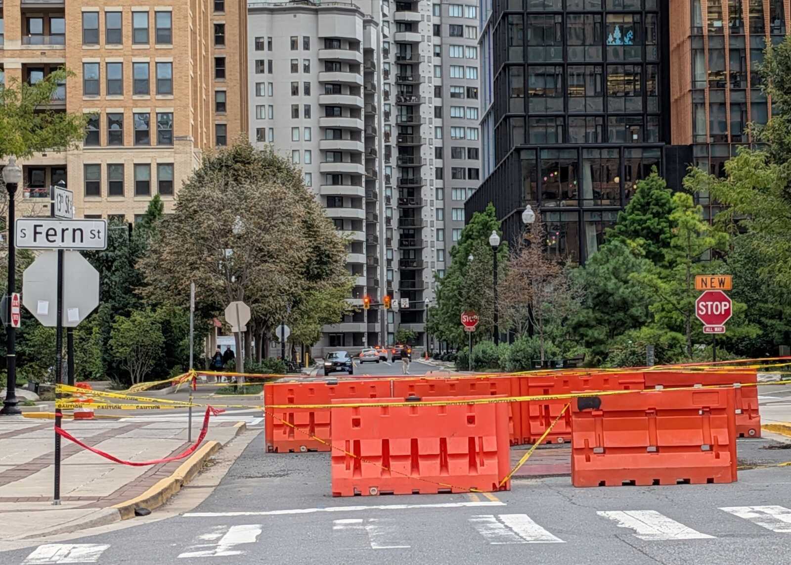 Urban street under construction with orange barriers and yellow caution tape across the road; high-rise buildings in background and a Stop sign on the right.