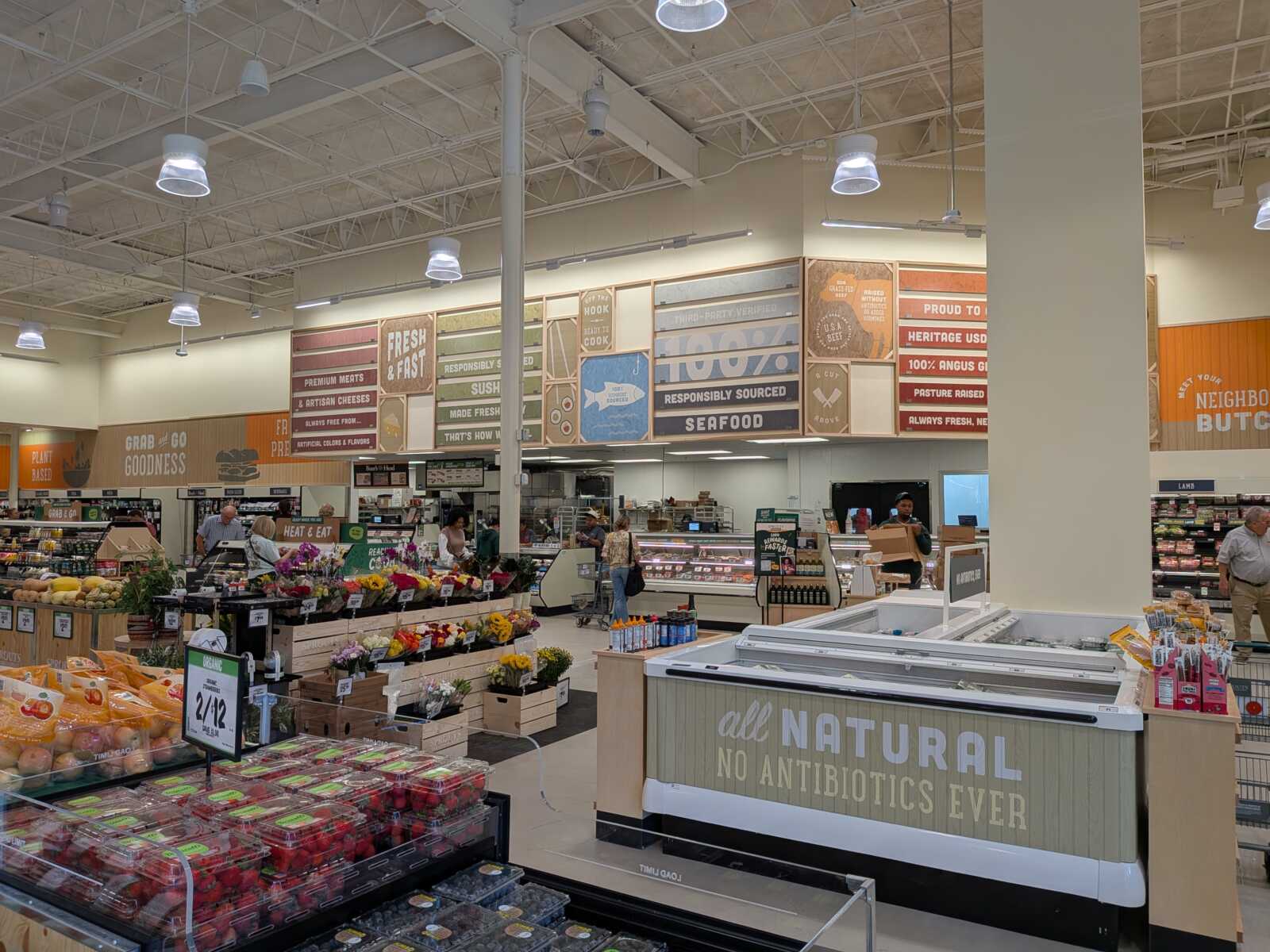 Wide view of a grocery store with produce displays, checkout counters, and signs overhead saying 'Natural No Antibiotics Ever'