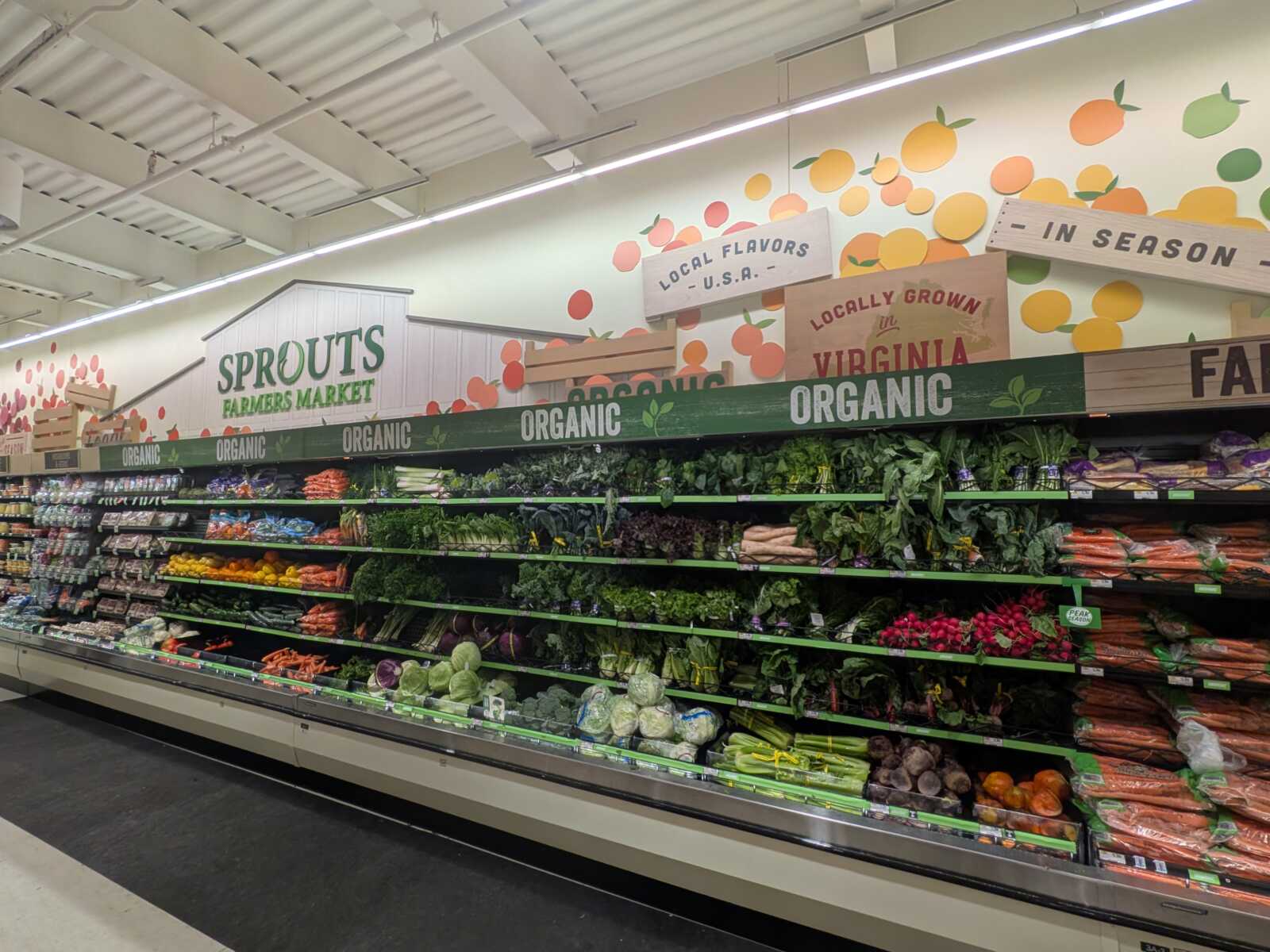 Produce aisle at Sprouts Farmers Market with organic signage and a colorful mural of fruit in the background, shelves stocked with greens, root vegetables, and colorful produce.