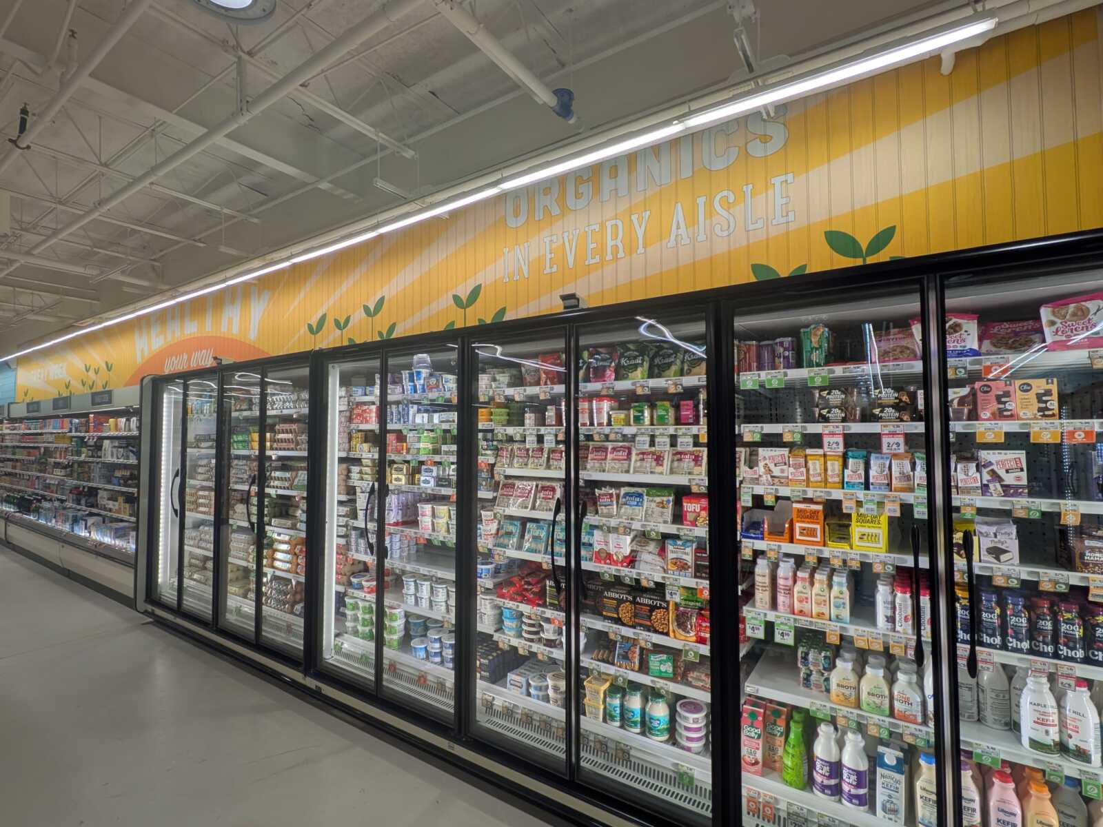 Grocery store dairy aisle with glass-door refrigerators and an orange sign reading “Organics in every aisle.”