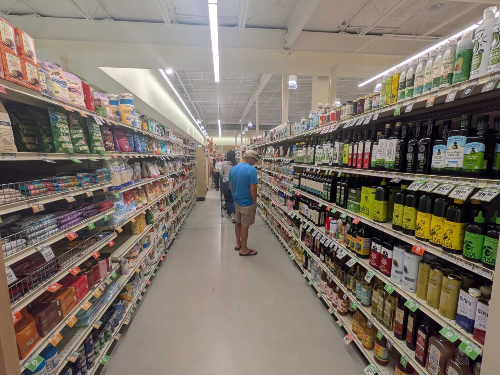 Man in a blue shirt and shorts stands in a grocery store aisle, facing away among tall shelves of bottles and packaged goods on both sides.