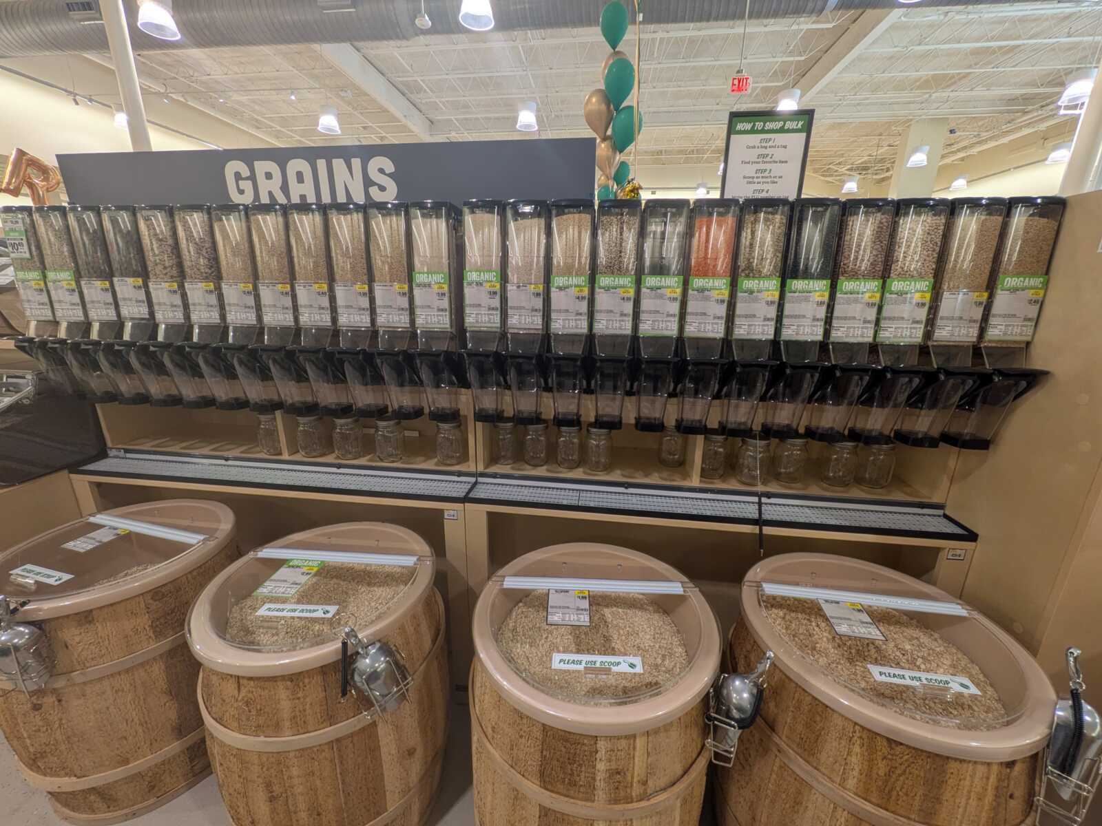 Row of bulk grain dispensers labeled Organic, mounted above wooden grain bins in a grocery store aisle marked 'GRAINS'.
