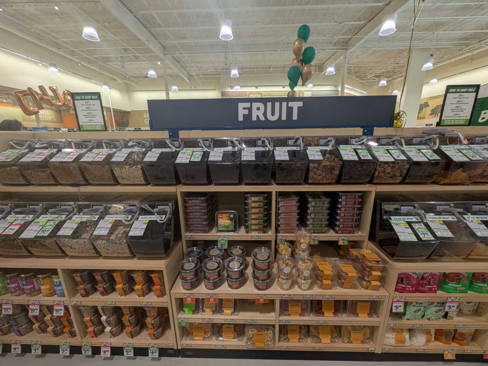 Bulk bins of nuts and dried fruits under a blue 'FRUIT' sign in a grocery store.