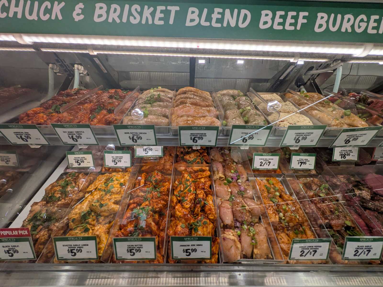Deli case with assorted marinated meats and kebabs in plastic trays, Sprouts signage above, price tags visible