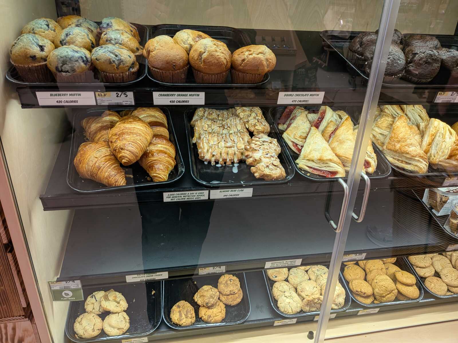 Shelf of baked goods: blueberry muffins and cranberry orange muffins on top row, double chocolate muffins to the right, all in trays inside a display case