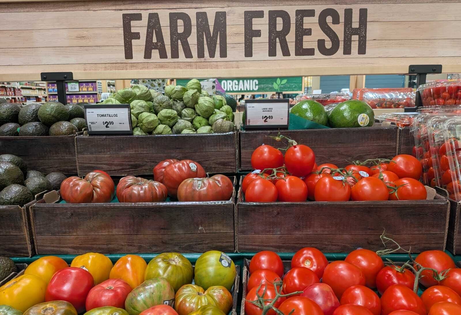 Wooden crates under a Farm Fresh sign display red tomatoes, green tomatoes, and avocados in a grocery store.