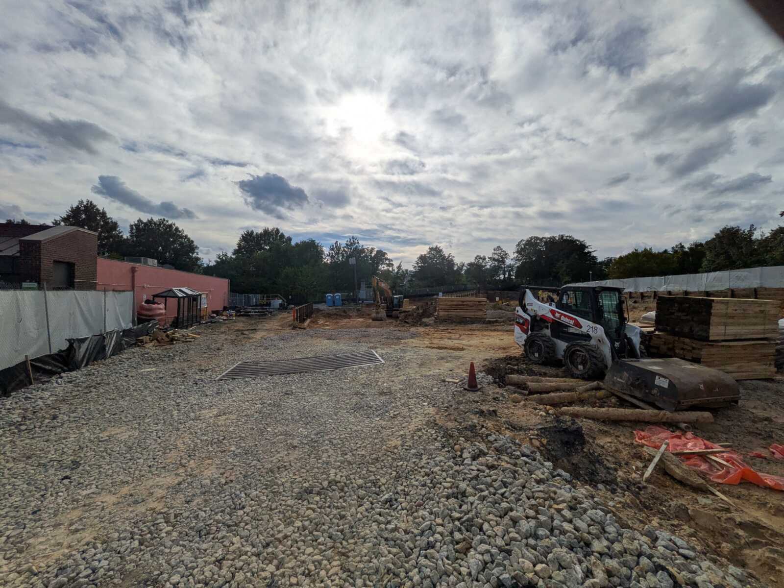 Construction site with a small skid-steer loader on the right, pallets of lumber, and a gravel yard under a cloudy sky