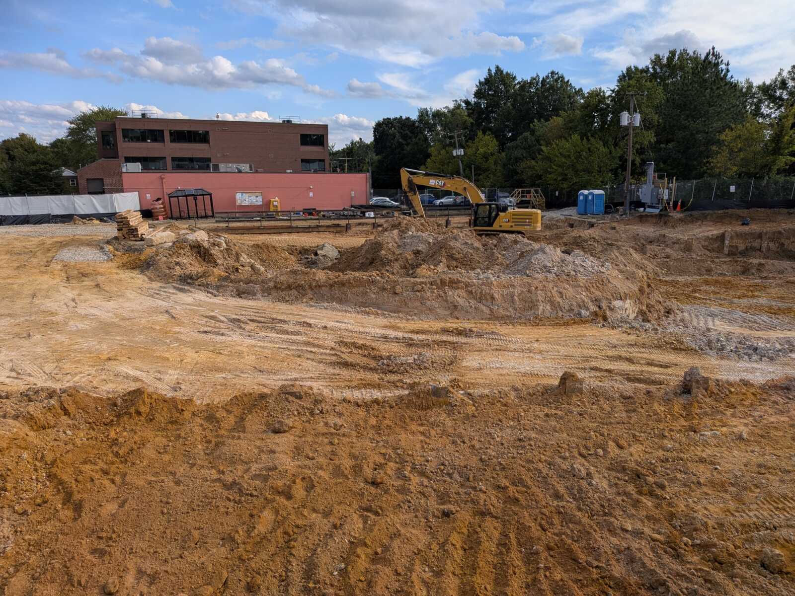 Construction site with a yellow excavator on a dirt mound and a red-brick building in the background