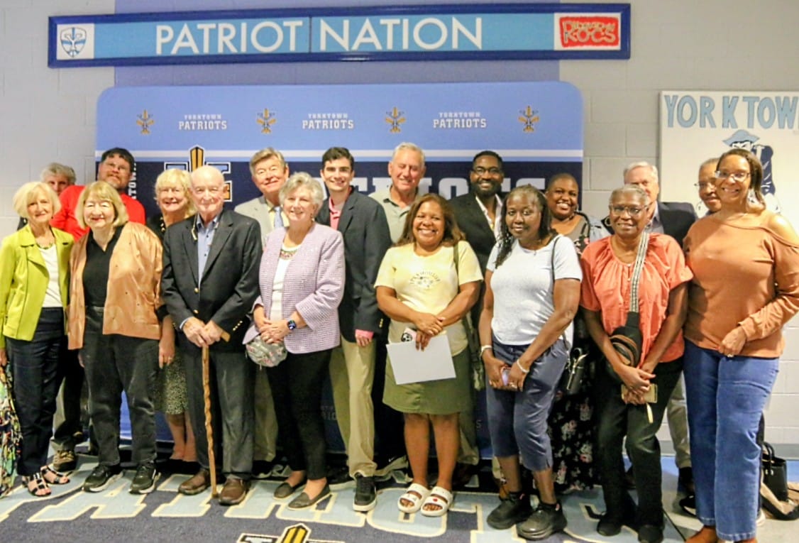 Group of diverse adults posing together in front of a'Patriot Nation' backdrop in a school or community center.