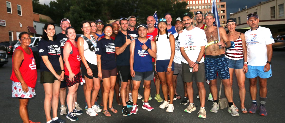 Group of runners posing for a post-race photo outdoors, wearing red, white, blue outfits and matching shirts; smiles and camaraderie evident with city buildings in background.