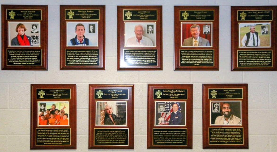 Nine wooden plaques mounted on a light brick wall, each plaque features a portrait photo and a block of biographical text honoring individuals.