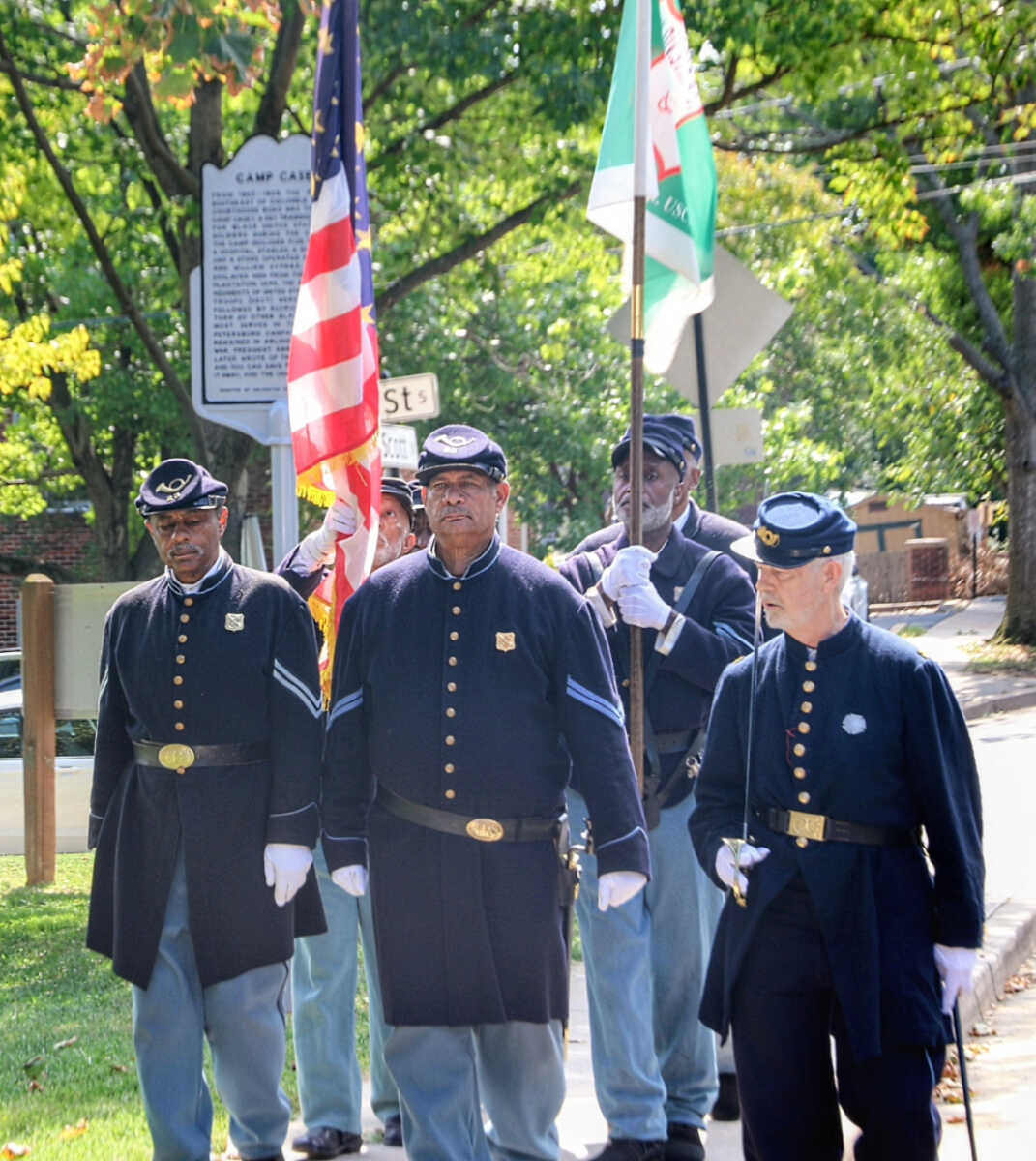 Group of men in historical navy-blue uniforms marching in a parade, carrying U.S. and flag banners.