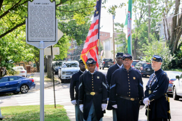 A group of uniformed veterans standing on a sidewalk beneath a historical marker that reads 'Camp Casey,' with American and other flags in the background.