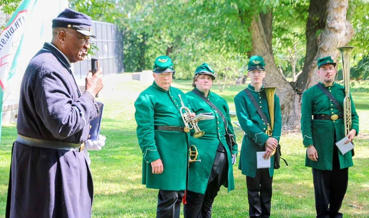Man in a dark uniform speaks into a microphone as four musicians in green coats with brass instruments stand in a park outside.
