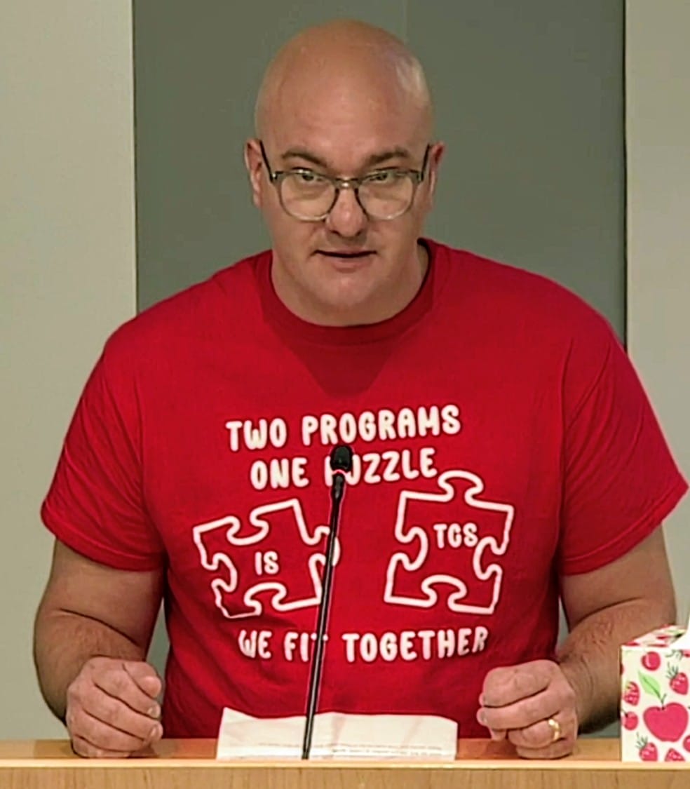 Robert Roberston speaks to the School Board about Integration Station (screenshot via APS) | ARLnow Bald man with glasses at a podium, wearing a red shirt with puzzle-piece graphics and text, speaking into a microphone.