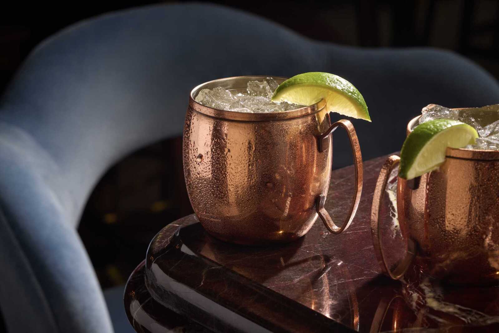 Copper mugs filled with crushed ice and lime wedges sit on a dark marble table in a moody lounge setting.