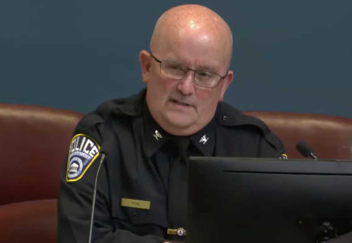 Police Chief Andy Penn at the County Board meeting (via Arlington County/YouTube) | ARLnow Bald police officer in a dark uniform with glasses sits at a desk, speaking into a microphone beside a computer monitor.